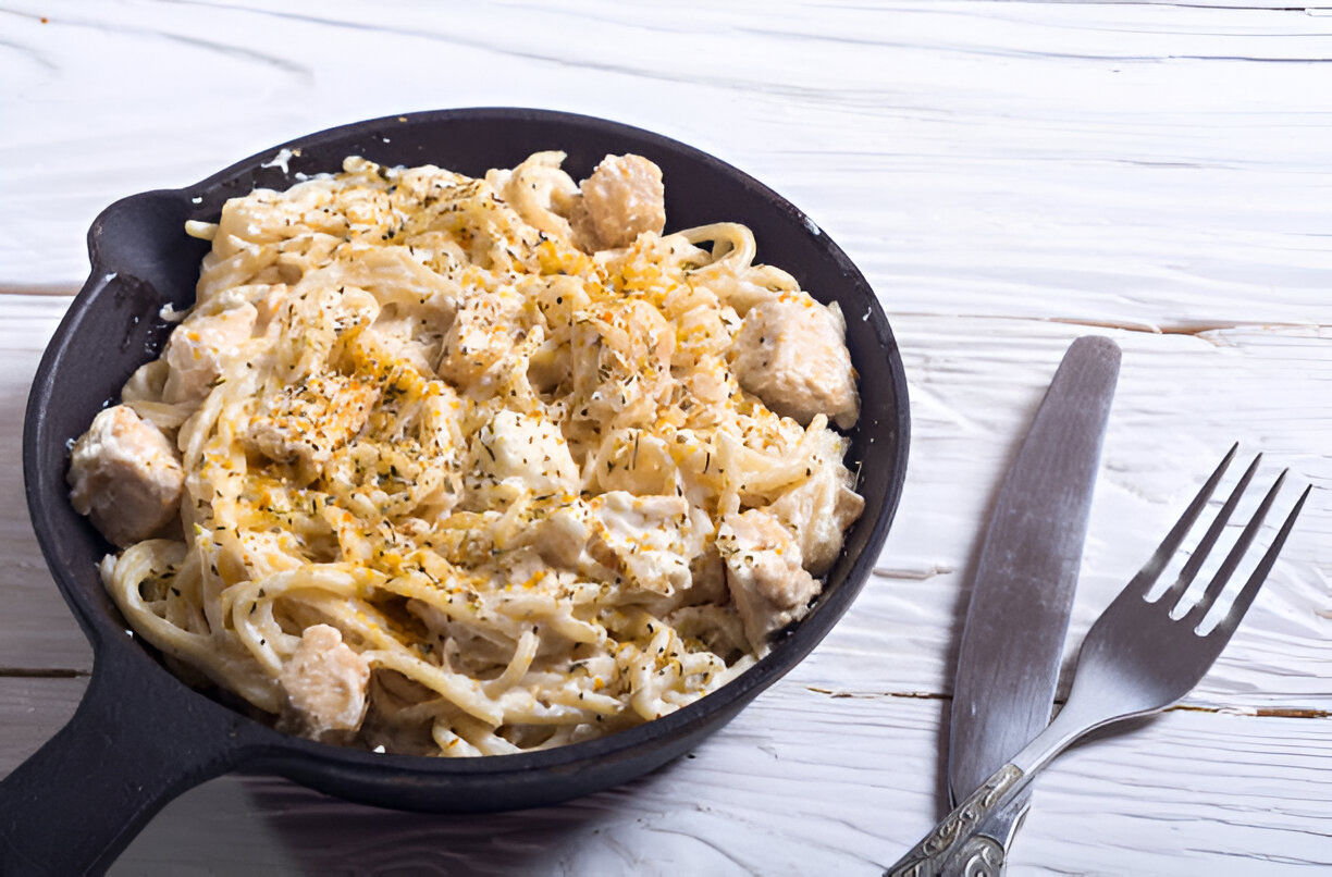 A skillet filled with creamy fettuccine pasta with chicken pieces, garnished with herbs, placed on a wooden surface.