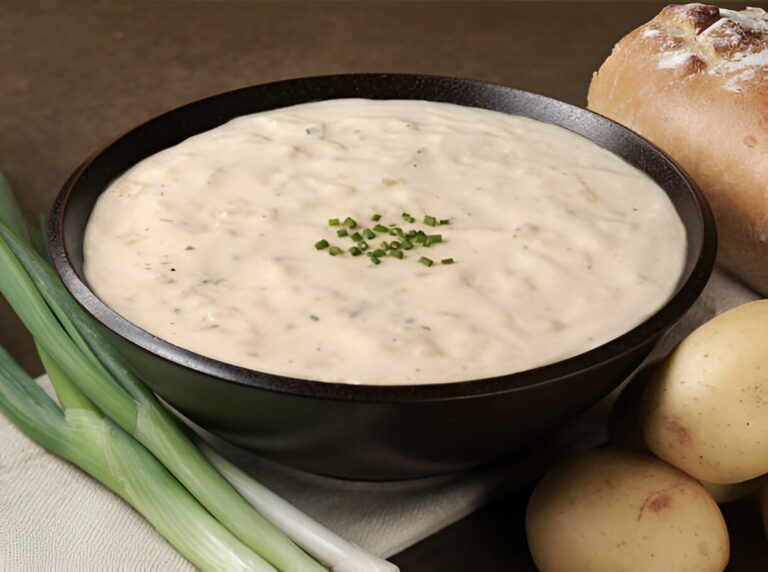 A bowl of creamy soup topped with chopped chives, accompanied by fresh green onions, potatoes, and a crusty bread roll.
