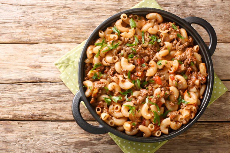 A delicious serving of macaroni pasta with ground beef, tomatoes, and green herbs in a black pot on a wooden table.