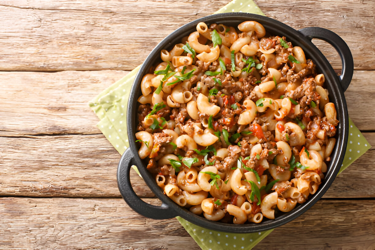 A delicious serving of macaroni pasta with ground beef, tomatoes, and green herbs in a black pot on a wooden table.