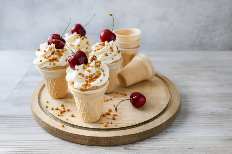 Four ice cream cones topped with whipped cream, cherries, and sprinkles on a wooden plate.
