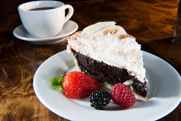 A slice of chocolate pie topped with whipped cream and accompanied by fresh berries, with a cup of coffee in the background.