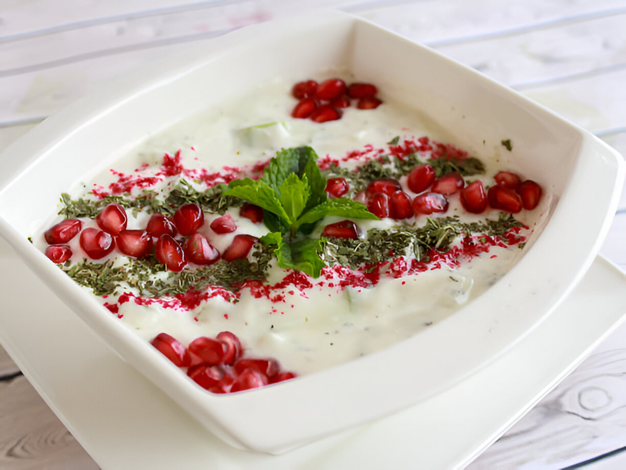 A bowl of yogurt dish garnished with pomegranate seeds, mint leaves, and herbs.