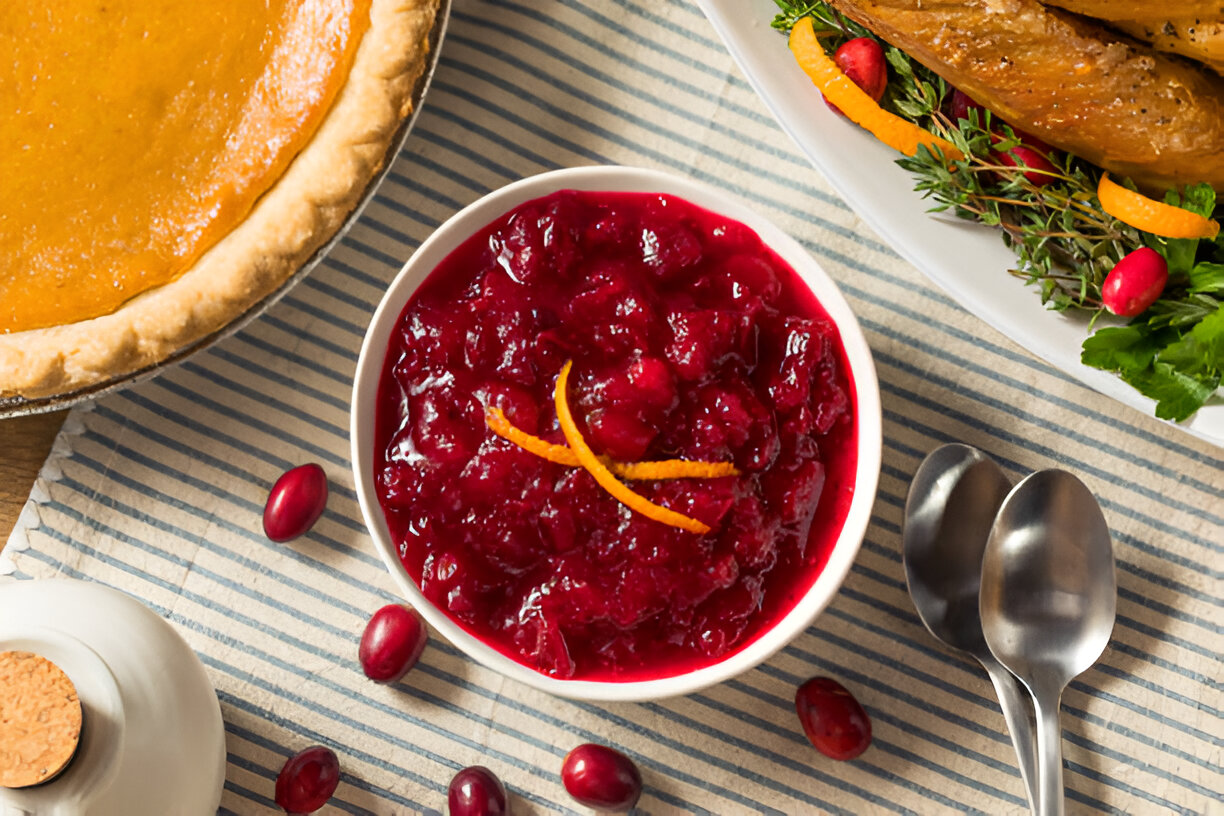 Cranberry sauce in a white bowl with orange peels on a striped tablecloth, surrounded by a pumpkin pie and garnished turkey