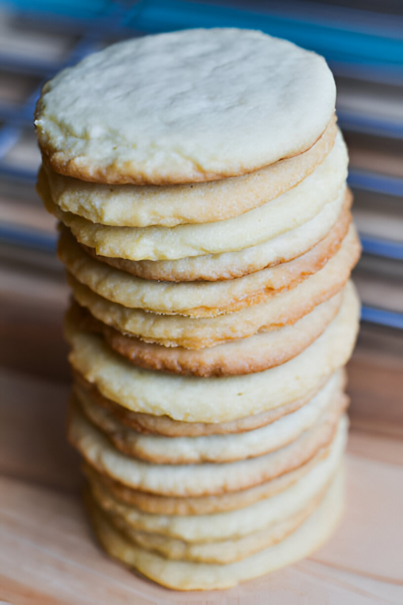 A tall stack of round, golden-brown cookies with a light, buttery texture on a wooden surface.