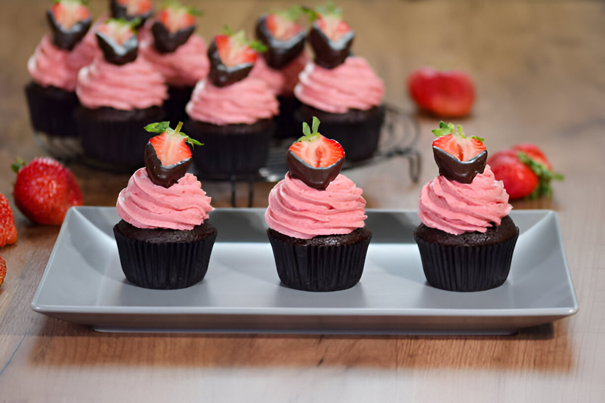 Three chocolate cupcakes with pink frosting and chocolate-covered strawberries on top, placed on a gray plate with fresh strawberries in the background.