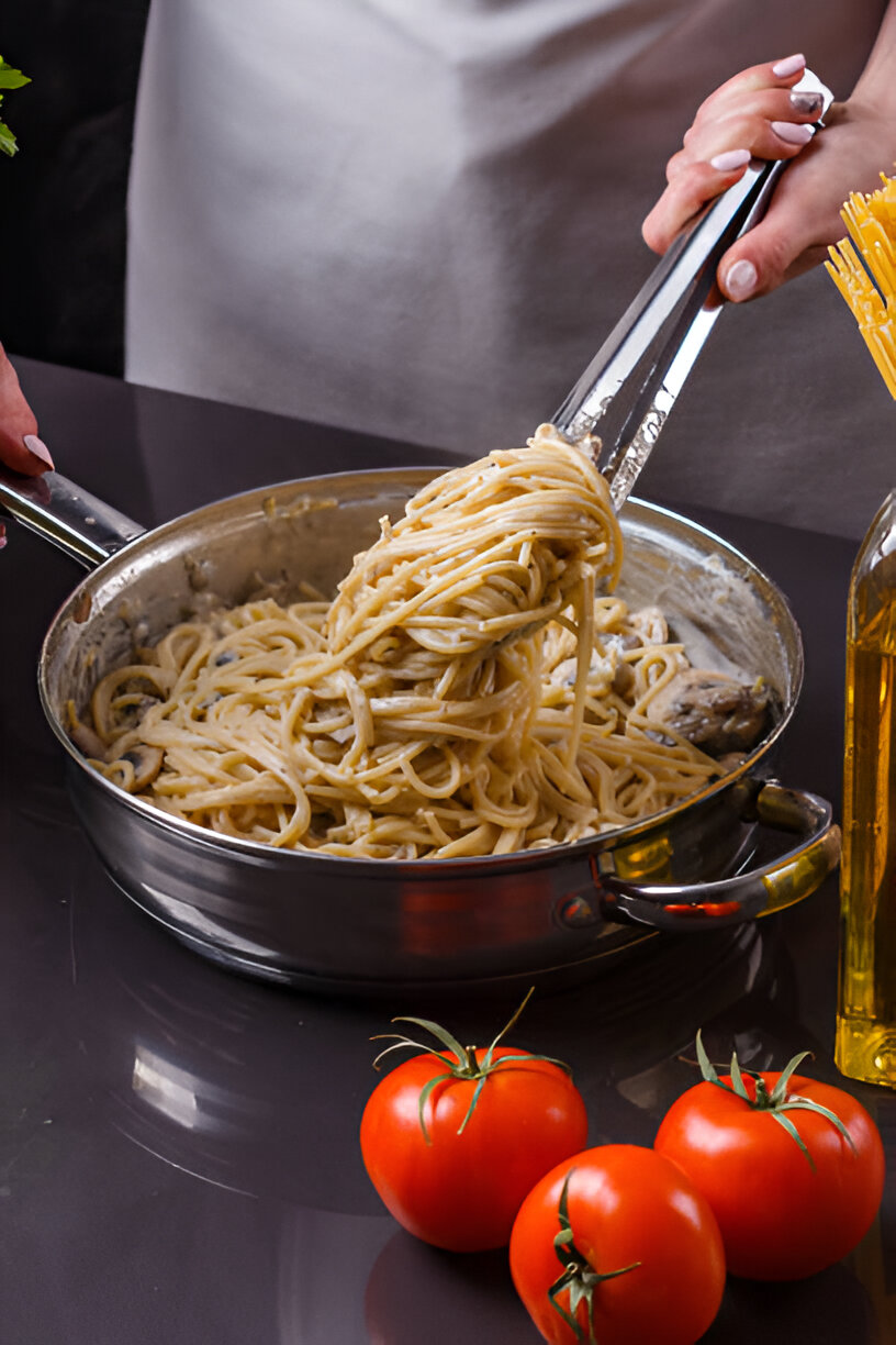 A chef using tongs to lift spaghetti from a stainless steel pan, with fresh tomatoes and olive oil in the foreground.