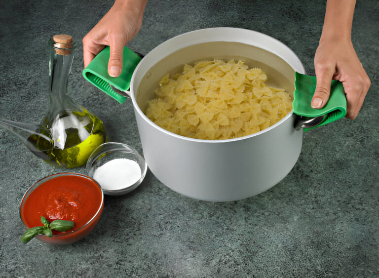 A person holding a pot of cooked pasta with a green silicone handle, surrounded by olive oil, tomato sauce, and salt.