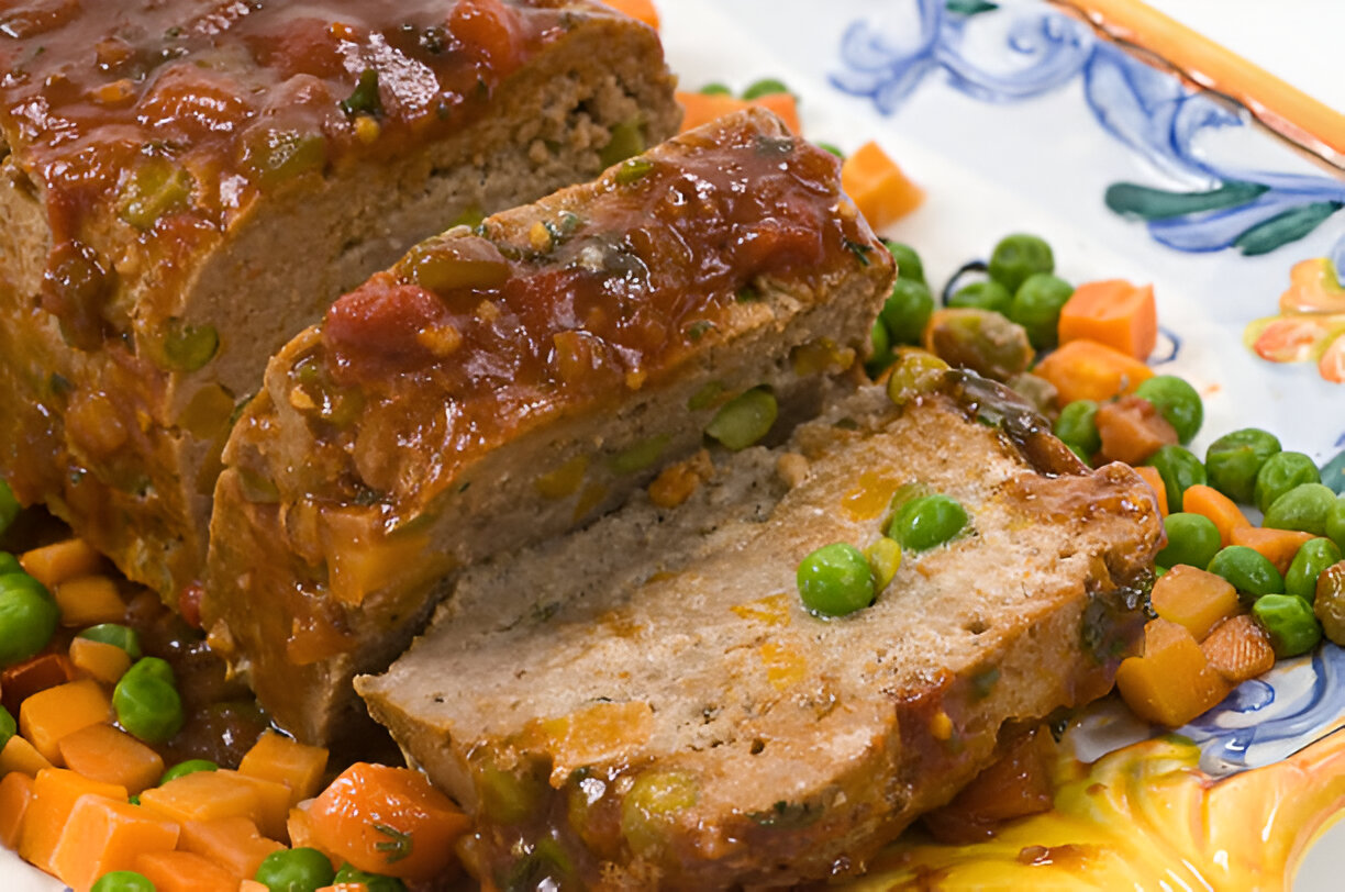 A delicious meatloaf topped with tomato sauce, surrounded by peas and carrots on a decorative plate.