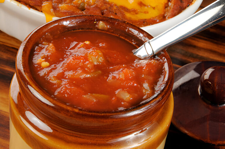A close-up view of a wooden bowl filled with salsa, featuring a silver spoon resting inside, with a casserole dish of cheese-topped food in the background.