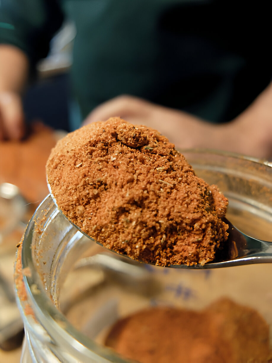 A close-up of a spoonful of spice being taken from a glass jar.