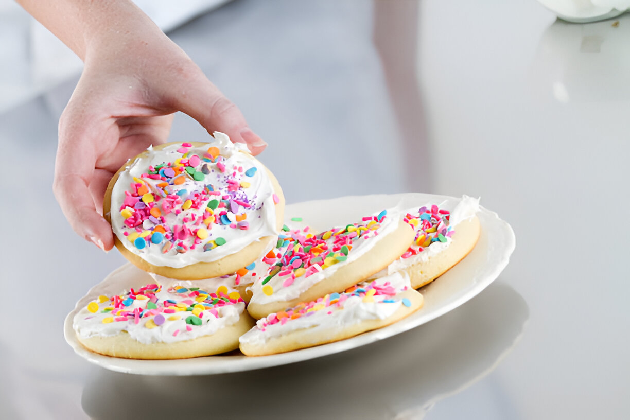 A hand holding a cookie topped with colorful sprinkles and frosting over a plate of other cookies.