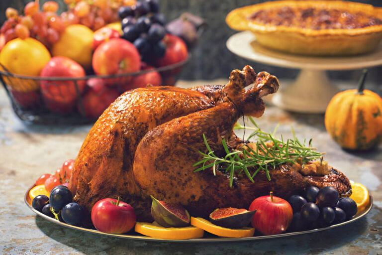 A beautifully roasted turkey garnished with rosemary, surrounded by a variety of colorful fruits including apples, grapes, and oranges, with a pumpkin and pie in the background.