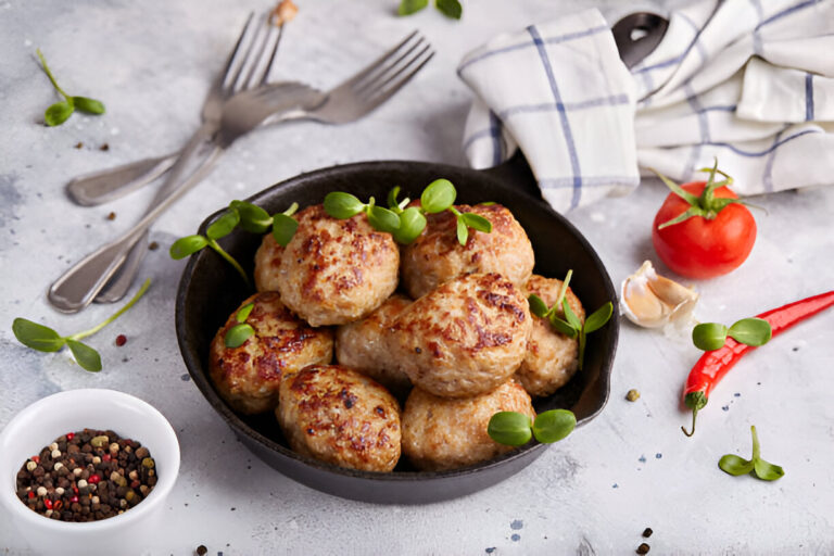 A black skillet filled with neatly arranged golden-brown meatballs garnished with fresh herbs, alongside spices, a tomato, and garlic.