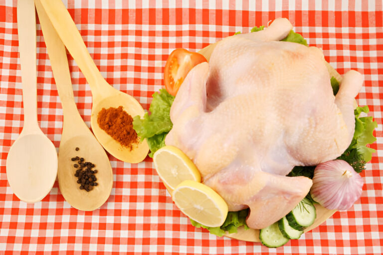 Raw whole chicken surrounded by fresh vegetables and spices on a red and white checkered tablecloth.
