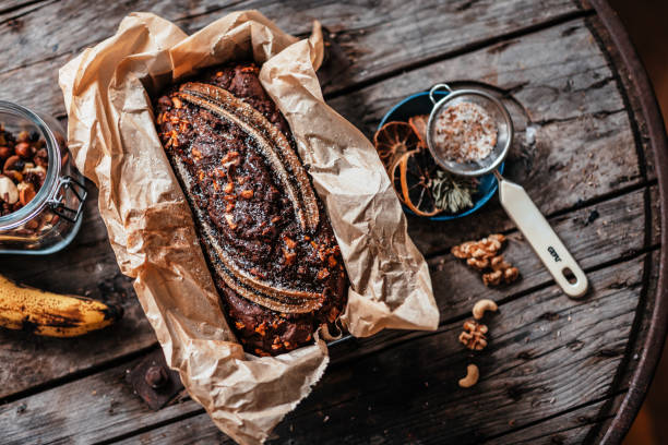 A freshly baked banana bread in a parchment-lined loaf pan, surrounded by nuts, a measuring spoon, and herbs on a rustic wooden table.