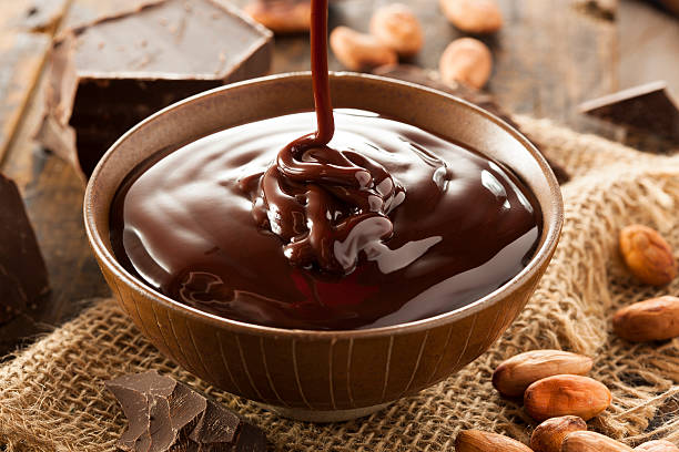 Bowl of rich chocolate sauce with a drizzle effect, surrounded by chocolate pieces and cocoa beans.