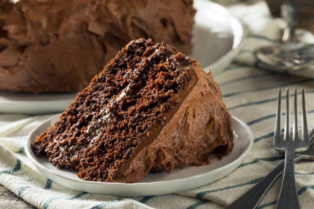 A slice of chocolate cake on a white plate, with a rich chocolate frosting, placed on a striped cloth