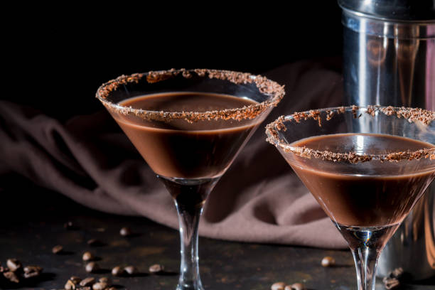 Two chocolate martinis with a chocolate rim, next to a shaker and coffee beans on a dark background.