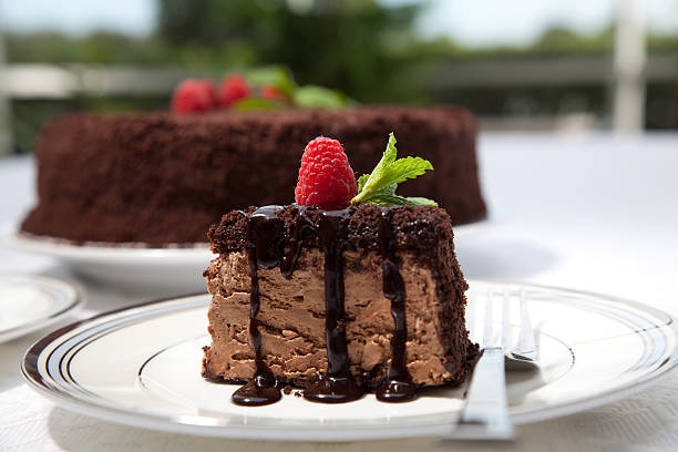 A slice of chocolate cake garnished with a raspberry and mint on a plate, with a whole cake in the background.