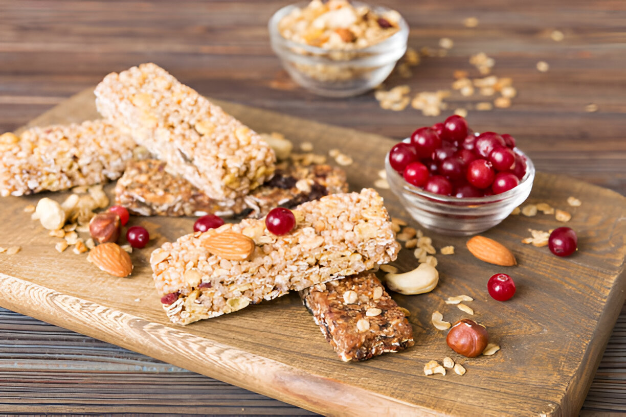A variety of granola bars and nuts on a wooden surface, accompanied by a bowl of fresh cranberries.