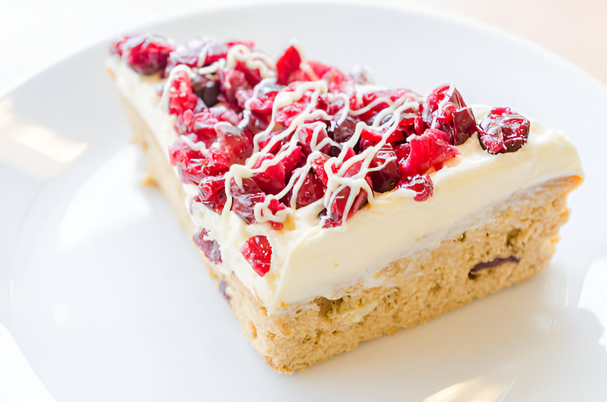 A slice of cheesecake topped with red cranberries and white chocolate drizzle on a white plate.