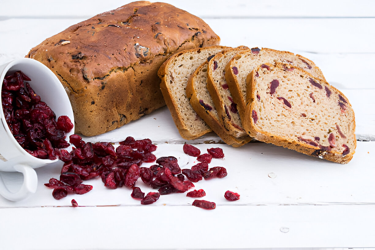 Sliced cranberry bread next to spilled dried cranberries on a wooden surface.