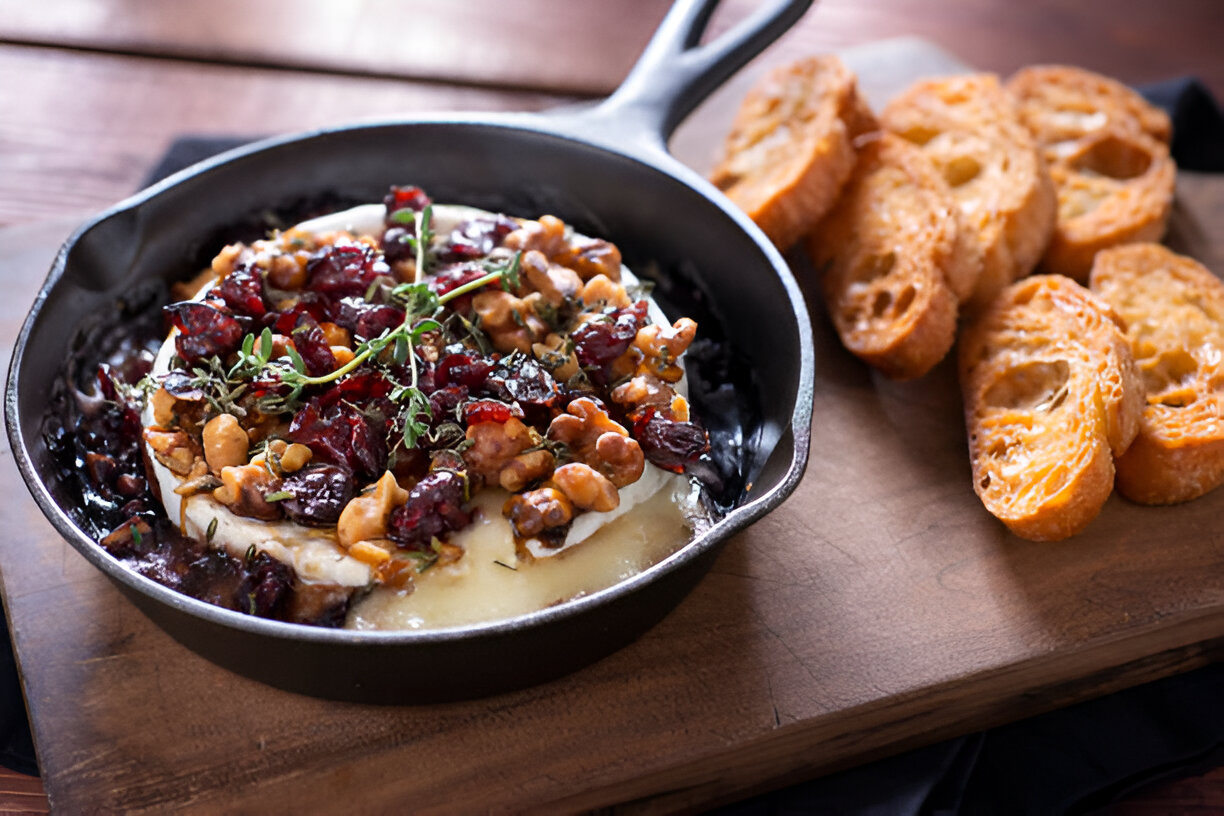 A cast iron skillet with baked cheese topped with nuts and cranberries, accompanied by slices of toasted bread on a wooden board.