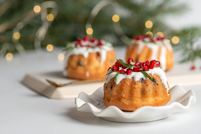 Mini bundt cakes topped with white icing, pomegranate seeds, and rosemary sprigs on a decorative plate, with festive lights in the background.