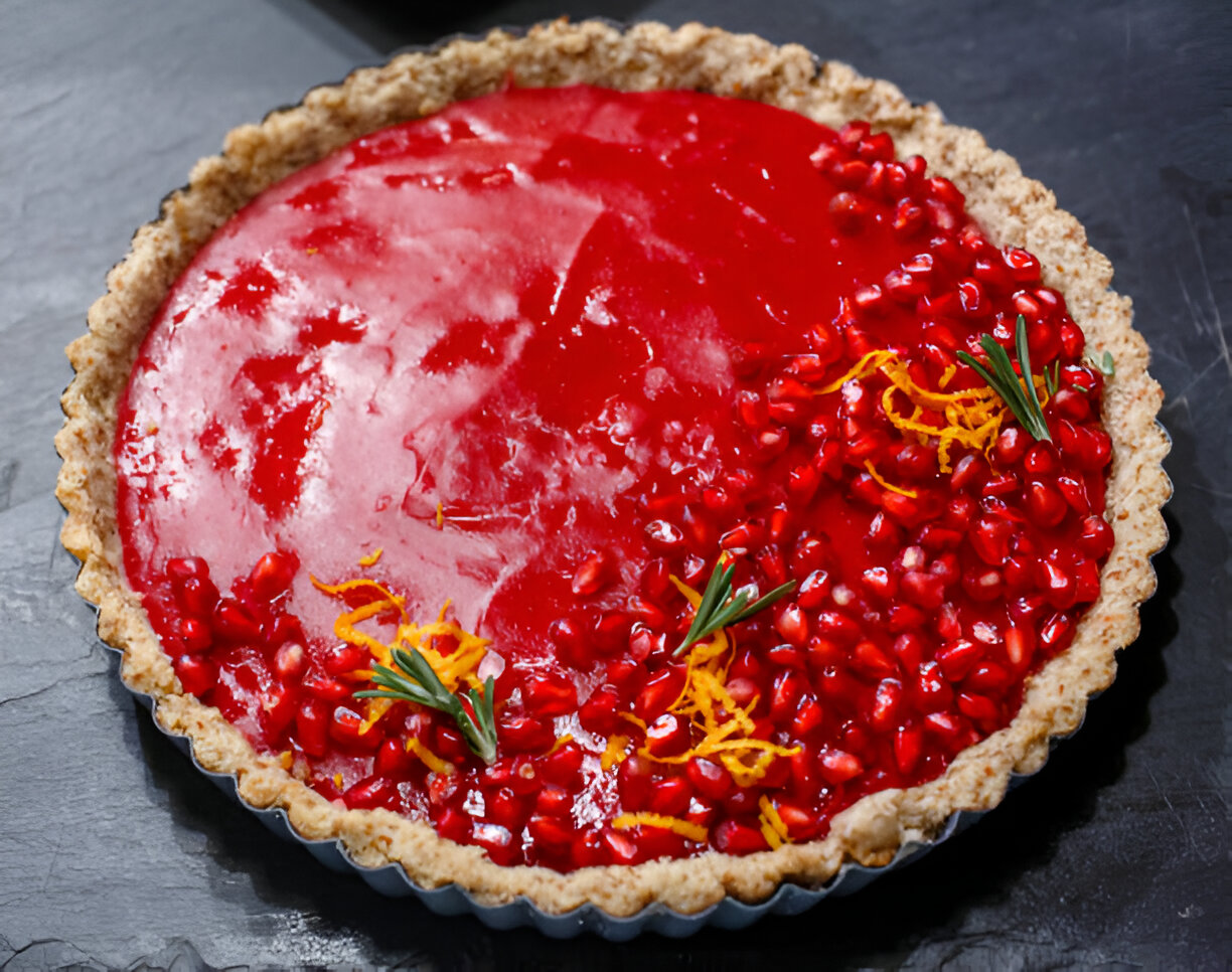 A vibrant pomegranate tart with a shiny red glaze, adorned with pomegranate seeds and orange zest, set on a black slate surface.