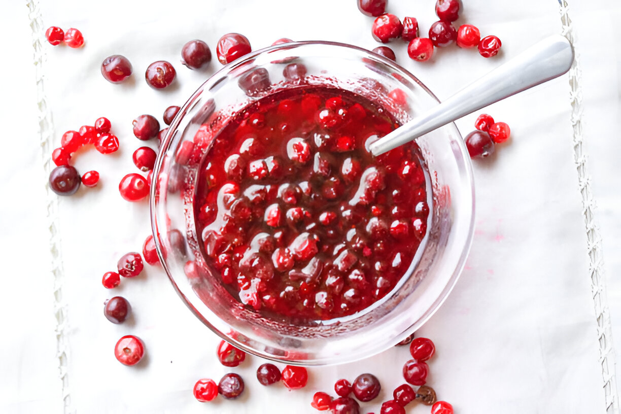 A clear bowl filled with cranberry sauce surrounded by fresh cranberries on a white surface.