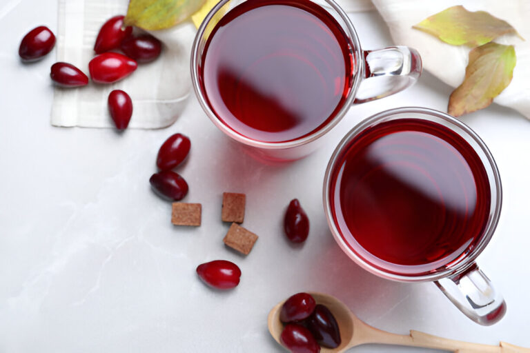 Two clear mugs filled with deep red liquid, surrounded by red berries and sugar cubes on a light surface.