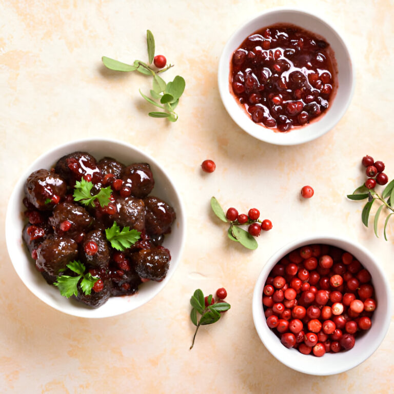 A top-down view of three white bowls containing dark brown meatballs with red berries, a berry sauce, and fresh red berries, surrounded by green herbs on a light background.