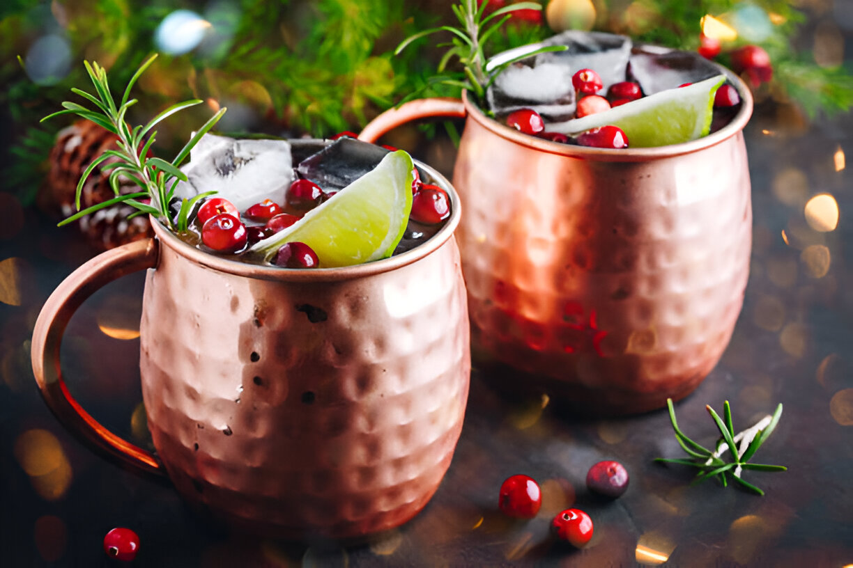 Two copper mugs filled with a vibrant cocktail, garnished with lime, cranberries, and rosemary, set against a blurred festive background.