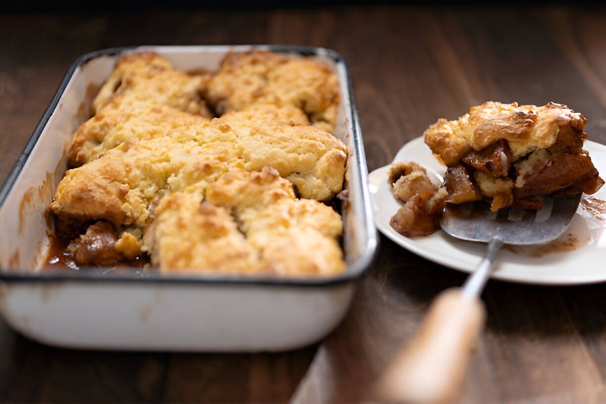 A freshly baked cobbler in a dish, served with a portion on a plate, featuring a golden-brown topping.