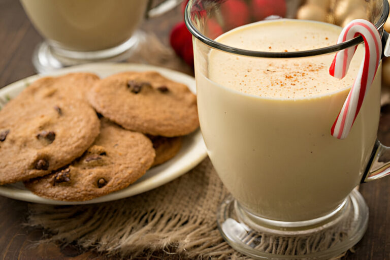 A glass of creamy eggnog with a candy cane stirrer beside a plate of chocolate chip cookies.
