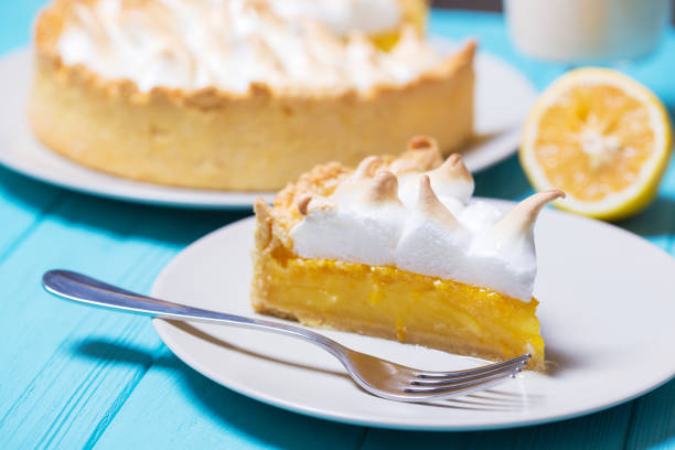 Slice of lemon meringue pie on a white plate with a fork, next to a whole pie and a lemon.