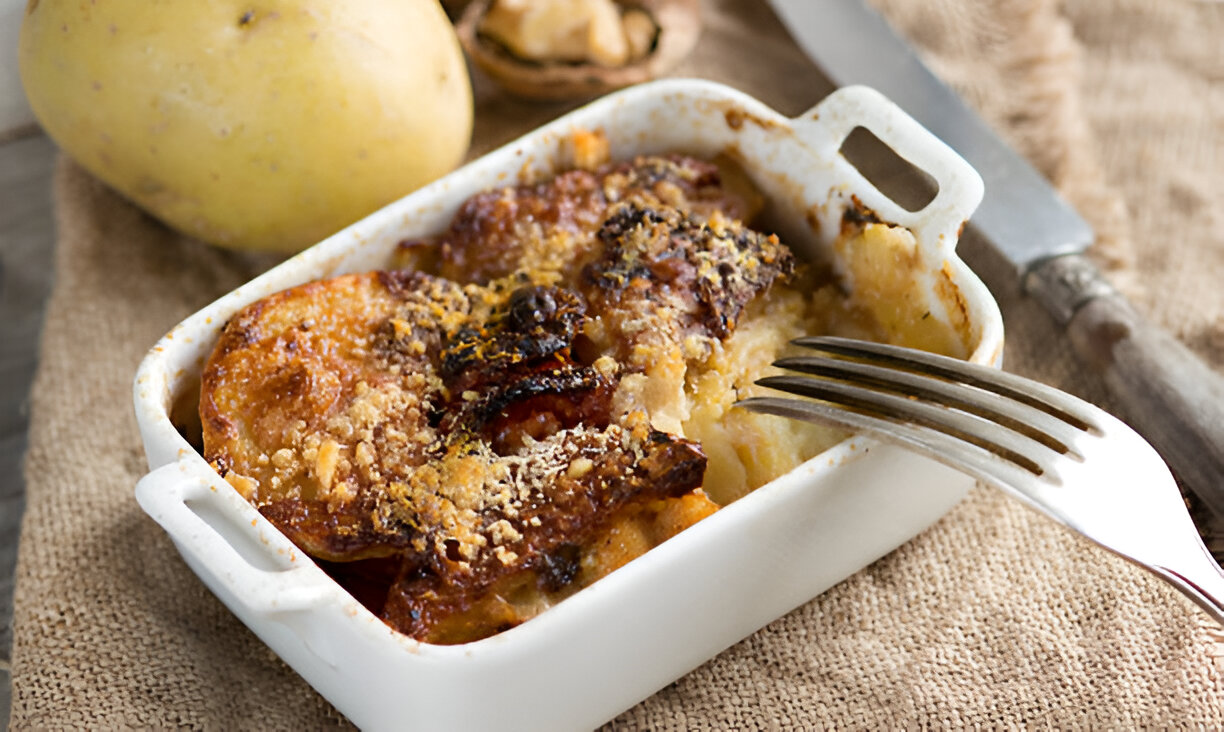 A baked potato dish with a crispy topping served in a small white baking dish, alongside a fresh potato and a fork.
