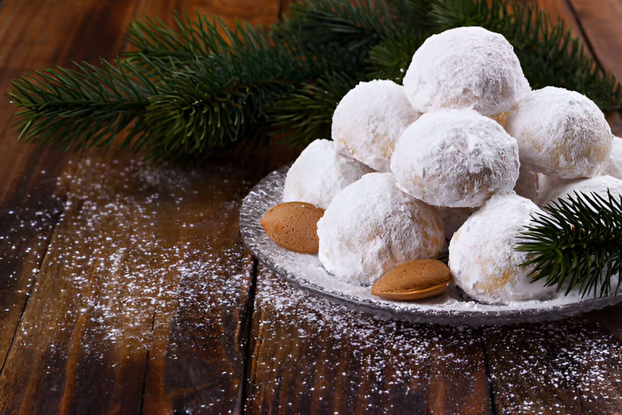 A platter of powdered sugar-dusted snowball cookies with almonds on a wooden table, accompanied by pine branches.