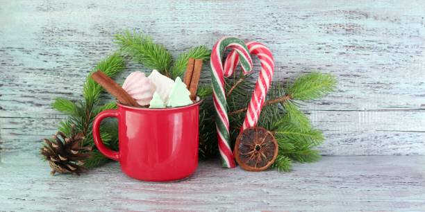 A red mug filled with marshmallows and garnished with candy canes and cinnamon sticks, surrounded by pine branches and a pine cone.