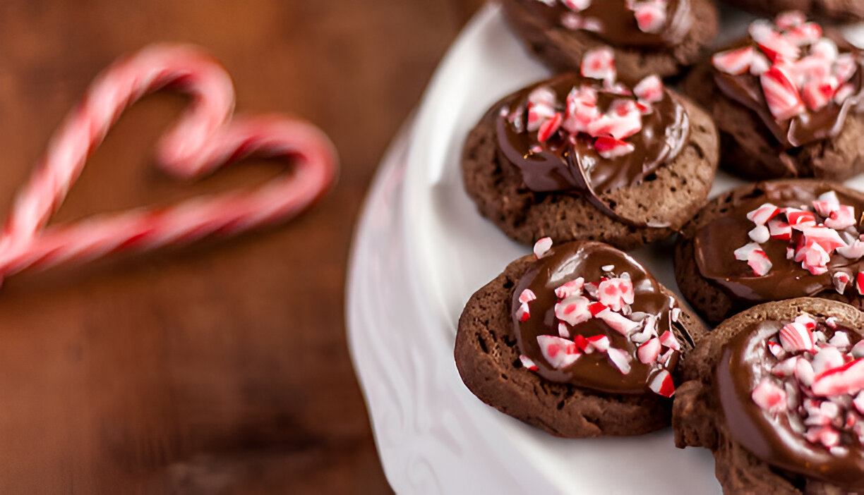 Close-up of chocolate cookies topped with crushed peppermint on a white plate, with a heart-shaped candy cane in the background.