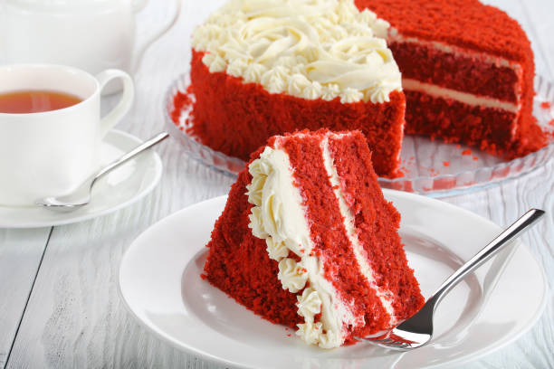 A slice of red velvet cake on a white plate with a fork, next to a cup of tea.