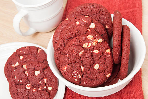 A bowl of red velvet cookies with white chocolate chips, with some cookies on a plate beside it and two white cups in the background.