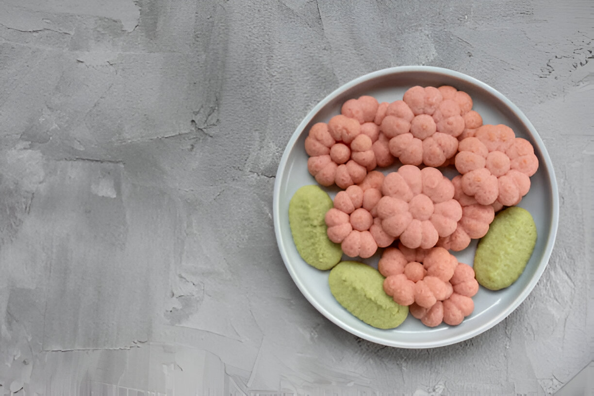 A plate of colorful snacks resembling flowers, featuring pink snacks shaped like blossoms and green oval snacks, on a textured gray background.