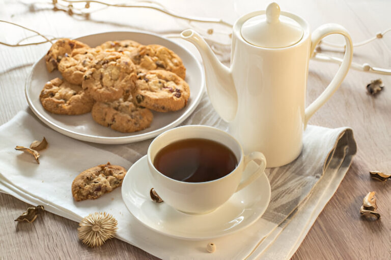 A cup of tea next to a teapot and a plate of cookies, with decorative natural elements on a wooden table.