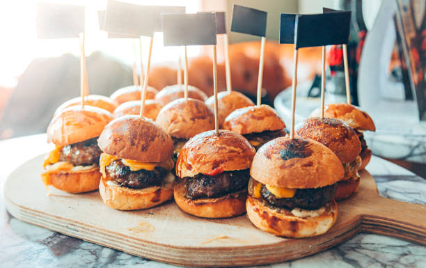 A wooden platter with several mini burgers topped with cheese and small black flags.