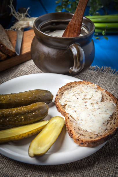 A plate featuring slices of bread with spread, accompanied by pickles and a small pot with a wooden spoon on a blue table.