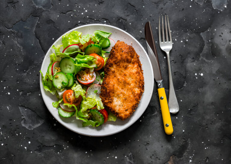 A plate of crispy breaded chicken paired with a fresh garden salad, featuring cucumbers, tomatoes, and lettuce, set against a dark stone background.