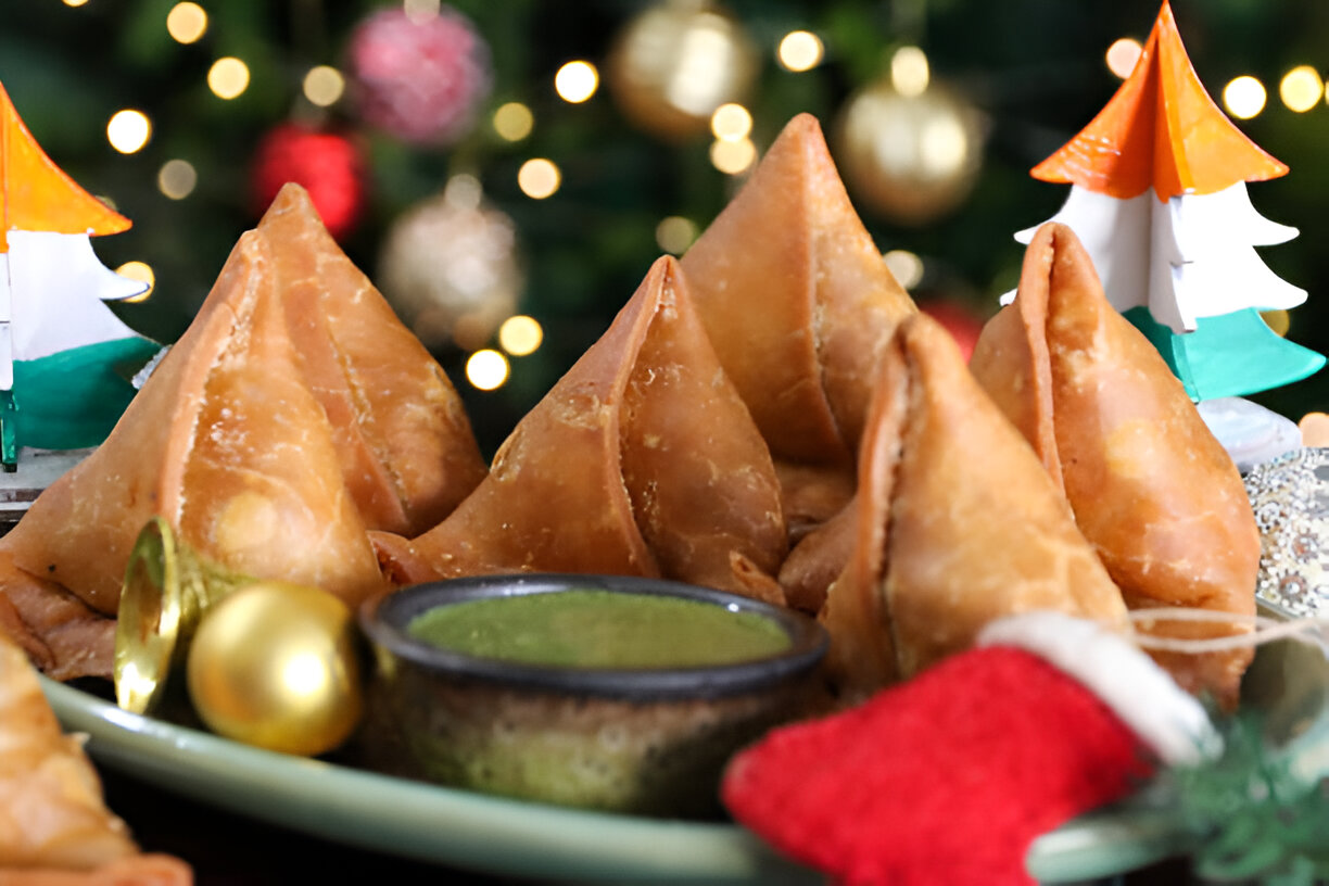 A plate of freshly fried samosas with a small bowl of green chutney, decorative holiday items in the background.