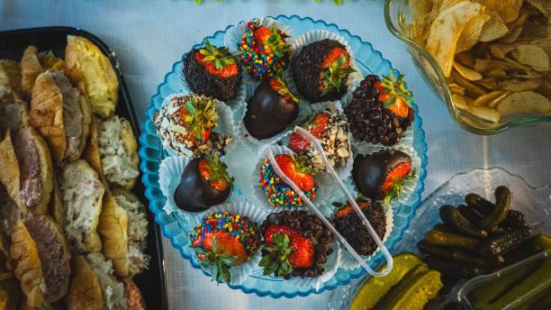 A colorful assortment of chocolate-covered strawberries arranged on a blue plate, surrounded by various snacks like chips and pickles.